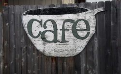 Cafe sign displaying weathered white paint with green letters in a cup shape, mounted on a rustic wooden fence