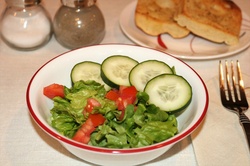 A fresh vegetable dinner salad in a white bowl, of lettuce, tomato and cucumbers, with garlic toast and condiments on a white table.